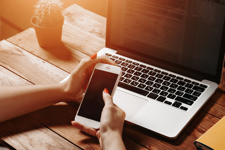 Female hands hold a smartphone near laptop and flower on vintage wooden table. Mock-up with laptop and smartphoneの写真素材