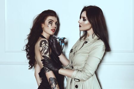 Beautiful Woman Tattoo Artist Does A Tattoo On The Girl's Hand With Creative Makeup. Fashion Photoshoot. Two Glamour Models Are Posing In Tattoo Studioの写真素材