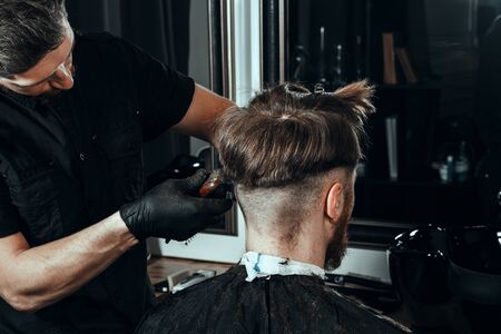 BARBERSHOP THEME. BEARDED SERIOUS BARBER IN BLACK RUBBER GLOVES IS TRIMMING THE HAIR OF HIS YOUNG HANDSOME CLIENT WITH CLOSED EYES. HE IS USING A HAIR CLIPPERの写真素材