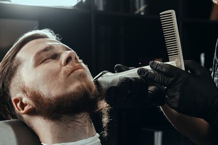 BARBERSHOP THEME. BARBER IN BLACK RUBBER GLOVES IS TRIMMING THE BEARD OF HIS YOUNG HANDSOME CLIENT WITH CLOSED EYES. HE IS USING A HAIR CLIPPERの写真素材