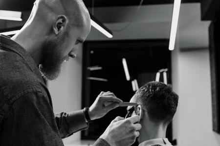 BARBERSHOP THEME. BALD BARBER IS TRIMMING THE HAIRCUT OF HIS BEARDED SERIOUS CLIENT. HE IS USING A...