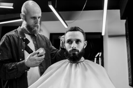 BARBERSHOP THEME. BALD BARBER IS TRIMMING THE HAIRCUT OF HIS BEARDED SERIOUS CLIENT. HE IS USING A HAIR CLIPPERの写真素材