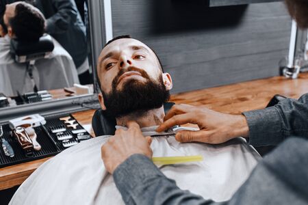 BARBERSHOP THEME. BALD BEARDED BARBER DRESSES A NEGLIGEE ON HIS YOUNG HANDSOME CLIENTの写真素材