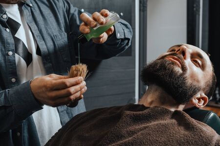 BARBERSHOP THEME. BALD BEARDED BARBER POURS A SHAVING-CREAM ON A BRUSH FOR HIS YOUNG HANDSOME CLIENTの写真素材