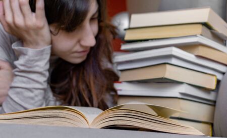 reading a novel on the sofa at home, next to a pile of books