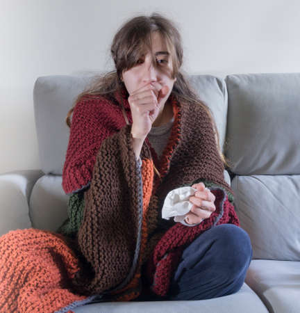girl sitting on the sofa, covered with a colored blanket, with the flu and with a tissue to blow her snotの写真素材
