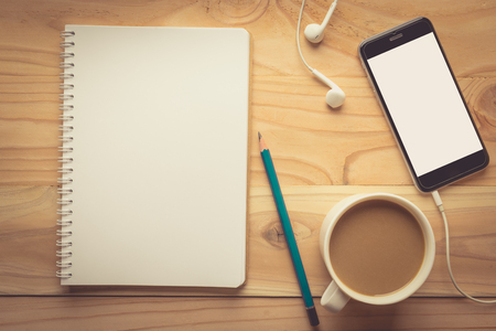 Top view of Smartphone with blank screen, earphones, notebook and coffee on wooden background , vintage toneの写真素材
