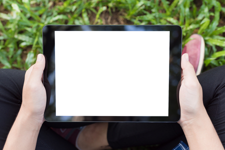 Mockup image of an asian woman sitting and holding black tablet with blank white screen on green grass at the parkの写真素材