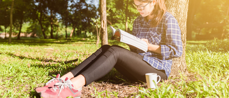 The girl sitting on a green grass with cup of coffee and reads the book, young woman  with coffee sitting on the grass and reading a book in the park banner panoramic crop for copy space.の写真素材