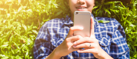 smiling young woman using and touching cell phone on meadow in park banner panoramic crop for copy space.の写真素材