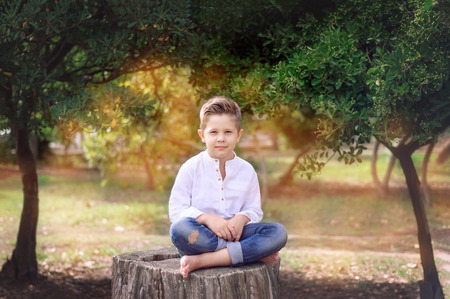 Boy 8 years old sitting on a tree stump on a sunny summer day. Kid outdoor enjoying nature. Handsome Boy barefoot sitting cross-legged.の写真素材