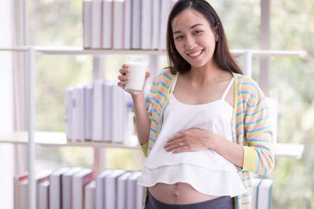 A pregnant woman is holding a glass of milk beside the window at home. Concept, Home activities and...