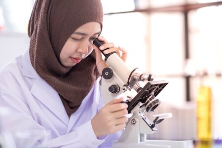beautiful female muslim scientist wearing white gown looking through microscope during experiment in chemical laboratoryの写真素材