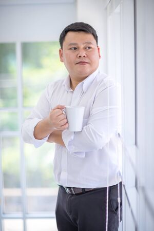 Asian Men Wearing a white shirt, standing and holding a white coffee cup. and look out the window confidently. Drink coffee, coffee shop, coffee break concept.の写真素材