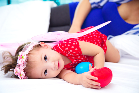 The cute asian girl wearing pink butterfly wing and lying on the white bed and playing with red heart, blurry mother pregnant background, happiness family conceptの写真素材