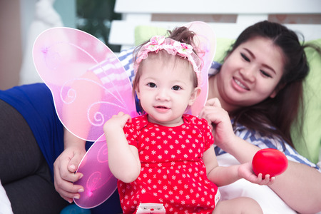 A little girl wearing pink butterfly wings is hold a red heart in the hand and playing with young mother on white bedの写真素材