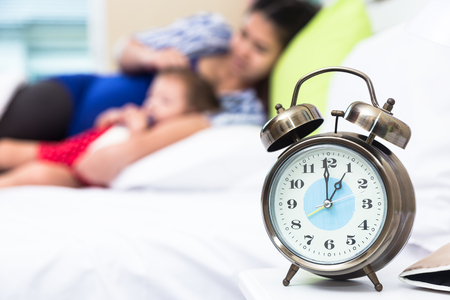 Selective focus a clock alarm with blurry image of mom and kid lying on the bed in sleep time during day, family conceptの写真素材