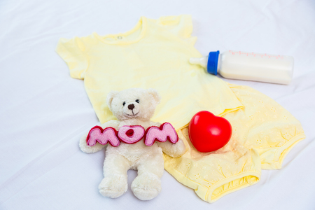 Selective focus at a teddy holding the word mom and red heart on the set of yellow clothing with baby milk bottle lying on white bedの写真素材