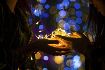 Close up the Hands of two female holding Yellow LED string light bulb within blurry bokeh at night, low lightの写真素材