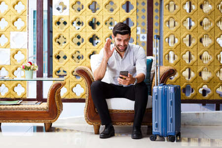 Young businessman sitting luggage and use smartphone video call in hotel corridor, copy spaceの写真素材