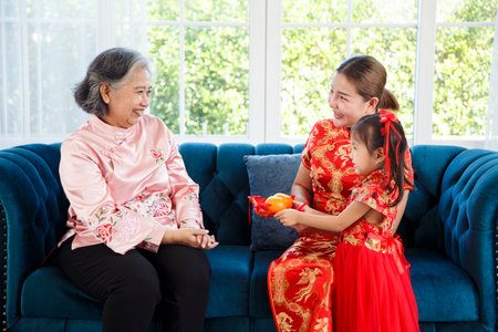 Asian young mother and daughter bring auspicious oranges to wish grandmother on Chinese New Year, They wearing chinese traditional clothing sitting on sofa at home, copy spaceの写真素材