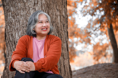 Portrait Beautiful senior woman sitting under big tree while relaxing in the park, looking forward...