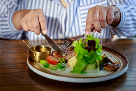 Close up and selective focus hands of asian Senior Woman use fork and knife while eating pork steak and Healthy Salad on dishの写真素材