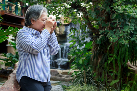 Asian senior woman farmer use digital camera to taking photo in beautiful garden, blurred artificial waterfall in the background, Relaxing time of retirement woman with happiness, copy spaceの写真素材