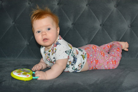 Red hair baby girl lying on a couch on a belly and holding a toy. 6 months baby girl having belly...