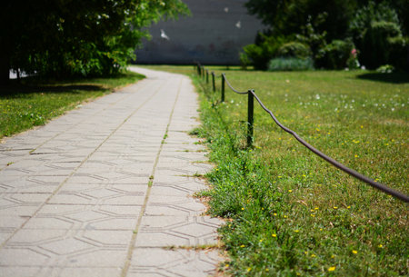 Footpath in a city with trees and metal wire fence at the sides. Sunny summer day. Tile pathway with blooming meadow in right side.の写真素材