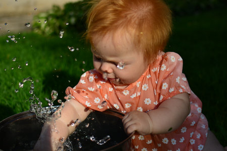Baby girl in pink dress is playing with water in pot in a back yard. Kid having fun outside in hot days.の写真素材