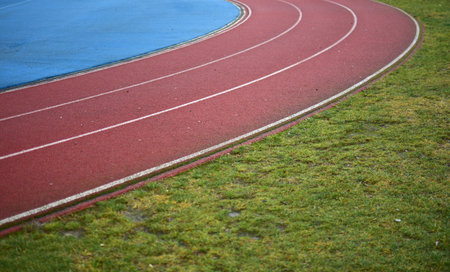 Red running track with blue sports field in left side and green grass in right side, sunny day