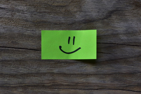 Smiley face on yellow sticky note on wooden board textured background. Black marker draw smiley face...