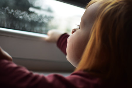 Red messy hair toddler girl curiously looking through the window. Daughter impatiently waiting parents.の写真素材