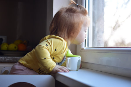 Red messy hair toddler girl curiously looking through the window. Daughter impatiently waiting parents.の写真素材