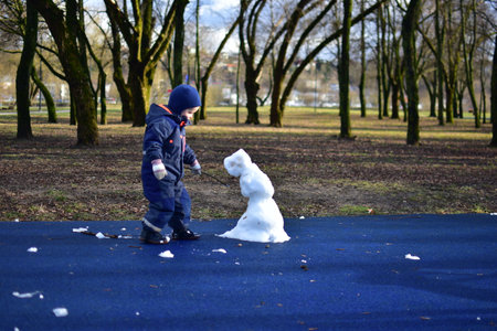 Toddler boy making snowman from the last snow in children playground in city park in spring. Kid and sad looking snowman waiting for winter end.の写真素材