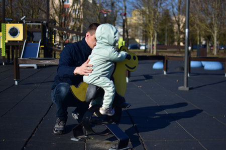 Toddler girl sitting on swing chair in playground in cold weather spring. Daddy holds daughter and helps to swing.の写真素材