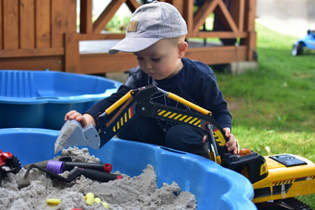 Toddler boy plays with toy excavator in a small sandbox in a back yard. Kid having fun outside in summer time.の写真素材
