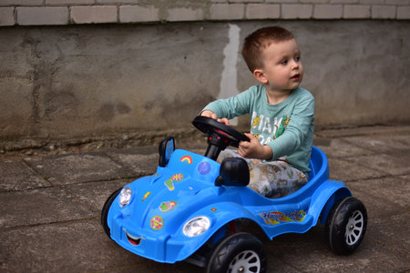 Toddler boy sitting in a big ride on toy car in a back yard. Kid driving toy push automobile outside in spring in europe.の写真素材