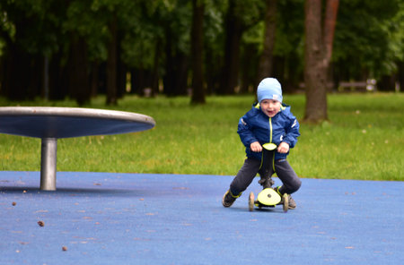 Happy toddler boy open mouth visible tongue sitting on green tricycle in public playground in spring. Little child riding a three wheel bike outdoors in cold spring.の写真素材