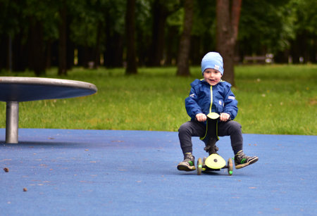 Happy toddler boy open mouth visible tongue sitting funny on green tricycle in public playground in spring. Little child riding a three wheel bike outdoors in cold spring.の写真素材