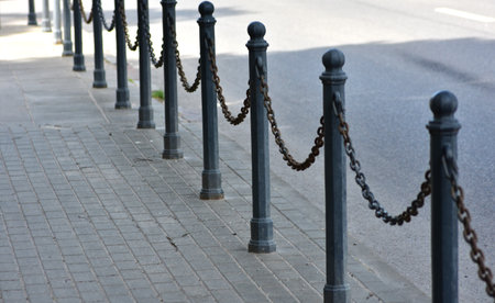 Iron posts with linked chains form a vintage street barrier along the sidewalk near a quiet road. Urban design element with perspective view, conveying order, boundary, and minimalism.の写真素材