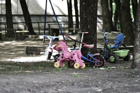 Abandoned playground with scattered children's tricycles and worn-out toys under trees. Dim light, moody tones, and silence evoke nostalgia, childhood memories, and a hint of forgotten joy.の写真素材