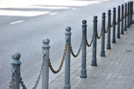 Iron posts with linked chains form a vintage street barrier along the sidewalk near a quiet road. Urban design element with perspective view, conveying order, boundary, and minimalism.の写真素材