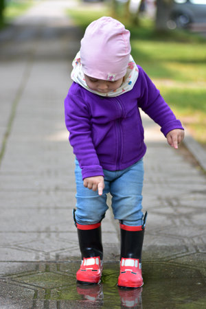 Cute toddler girl in red rain boots and colorful clothes playing in a puddle on a sidewalk during a sunny day. Childhood joy and outdoor fun after rain.の写真素材