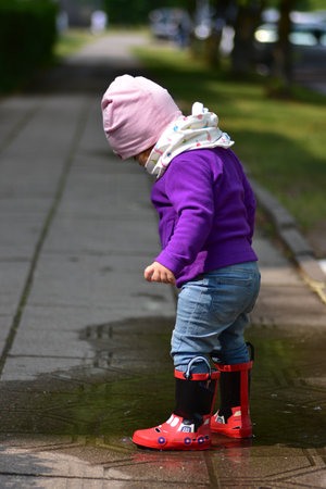 Cute toddler girl in red rain boots and colorful clothes playing in a puddle on a sidewalk during a sunny day. Childhood joy and outdoor fun after rain.の写真素材