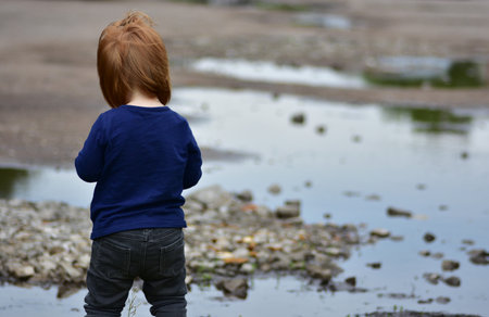 Red-haired toddler girl in dark clothes stands near puddles and rocky ground. Scene gives a moody, somber atmosphere. A candid, expressive view of childhood in raw outdoor environment.の写真素材