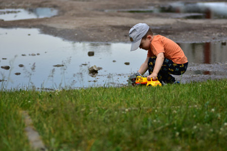 Young boy in cap and bright clothes plays with yellow toy truck near a puddle on grass. Child is focused and engaged. Candid outdoor moment showing creativity and exploration in nature.の写真素材