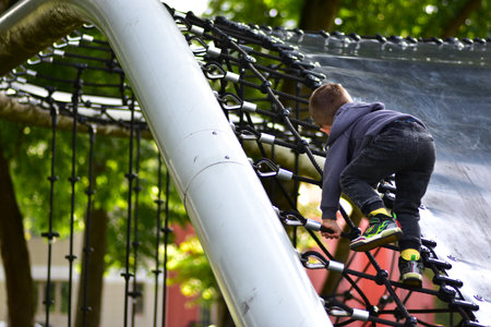 Little toddler boy in a hoodie climbs rope net at outdoor playground. Concentrated toddler enjoying adventure and physical activity in park. Child development and motor skills in action.の写真素材