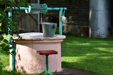 Old countryside water well with a green metal crank and a silver bucket. Surrounded by green grass and garden elements. Vintage rural equipment on a bright sunny day.の写真素材